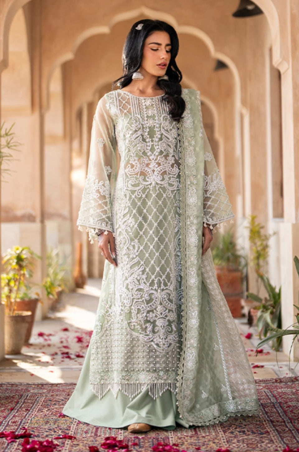 Woman in a light green embroidered dress standing in an ornate indoor setting with arches and plants.